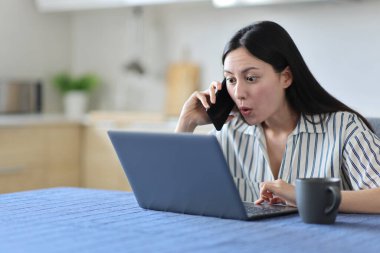 Şaşırmış Asyalı kadın telefonda konuşuyor. Evdeki mutfakta dizüstü bilgisayarını kontrol ediyor.