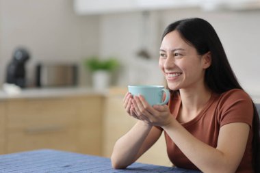 Mutlu Asyalı kadın başka tarafa bakıyor. Kahve içiyor. Evdeki mutfakta oturuyor.