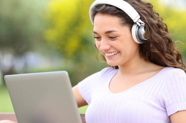 Parkta oturan, kulaklık takan, dizüstü bilgisayar kullanan mutlu kadın.