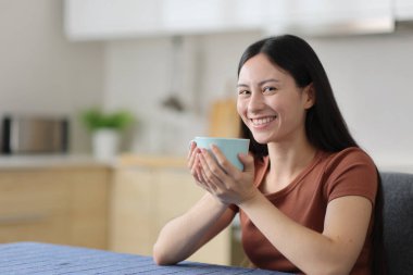 Mutlu Asyalı kadın kahve içiyor. Evdeki mutfakta sana bakıyor.