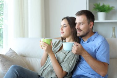 Rahatlamış, ciddi bir çift kahve içiyor ve pencereden dışarı bakıyor. Evdeki koltukta oturuyor.