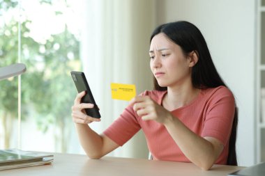 Üzgün Asyalı kadın internette telefon ve kredi kartıyla bir odada oturuyor.