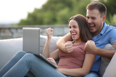 Heyecanlı çift dizüstü bilgisayarın içeriğini kontrol ediyor ve evde bir terasta kutlama yapıyor.