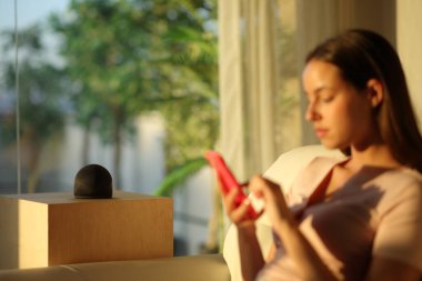 Gün batımında akıllı bir hoparlörün yanında akıllı telefon kullanan bir kadın.