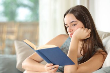 Evde bir kanepede oturmuş hayal kırıklığı yaratan bir kağıt kitap okuyan sıkıcı bir kadın.