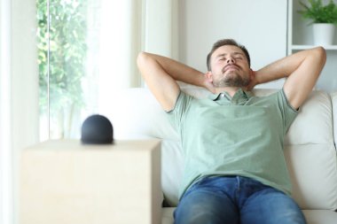 Evdeki kanepede oturan akıllı bir hoparlörün yanında dinlenen adam.