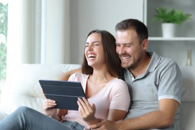 Komik çift tabletten medya içeriğini izliyor ve evde bir kanepede oturmuş kahkahalar atıyorlar.