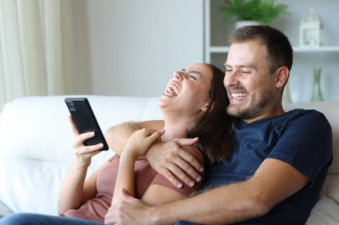 Komik çift birlikte gülüyor evdeki koltukta oturan telefon içeriğini kontrol ediyor.