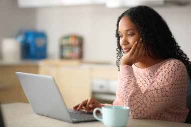Evdeki bir mutfakta dizüstü bilgisayar kullanan siyahi bir kadın.