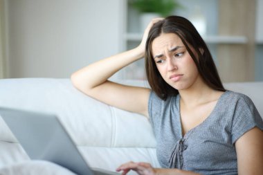 Kafası karışmış bir kadın dizüstü bilgisayarın içeriğini kontrol ediyor. Evin içindeki bir koltukta oturuyor.