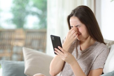 Cep telefonu kullanan bir kadın evdeki koltukta otururken göz yorgunluğu çekiyor.
