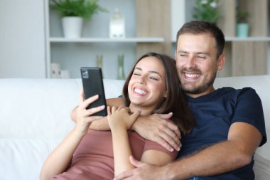 Mutlu çift cep telefonunu kontrol ediyor ve gülüyor. Evdeki koltukta oturuyor.
