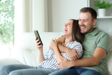 Mutlu çift yüksek sesle gülüyor. Cep telefonunun içeriğini kontrol ediyor. Evdeki koltukta oturuyor.