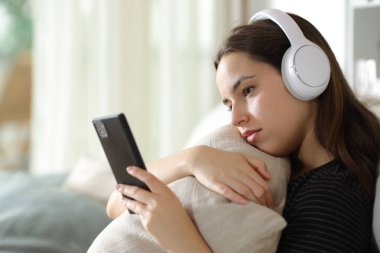 Kulaklık takan, cep telefonunu kontrol eden, evdeki koltukta oturan ciddi bir kadın.