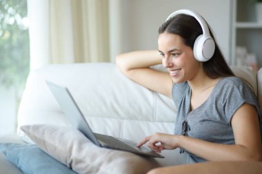 Kulaklık takan mutlu bir kadın dizüstü bilgisayarda eğlence içeriği seyrediyor evdeki bir koltukta oturuyor.