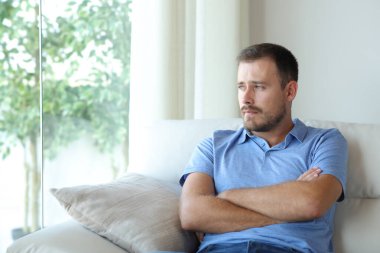 Evdeki kanepede oturup pencereden bakan dalgın üzgün bir adam.