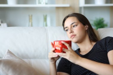 Ciddi düşünceli kadın kahve içiyor. Evdeki kanepede oturuyor.