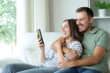 Mutlu çift cep telefonuyla medyayı izlemekten zevk alıyor. Evdeki koltukta oturuyorlar.