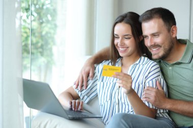 İnternetten kart ve dizüstü bilgisayarla ev içi bir koltukta oturan mutlu çift.