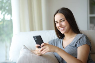 Cep telefonuyla poz veren mutlu kadın evdeki koltukta oturuyor.