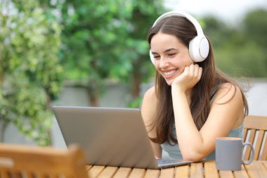 Evdeki terasta oturup bilgisayar ve kulaklık kullanarak medyayı izleyen mutlu kadın.