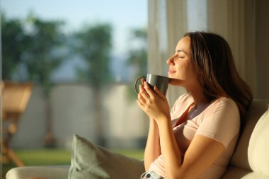 Mutlu kadın gün batımında kahve kokusu alıyor. Evdeki kanepede oturuyor.