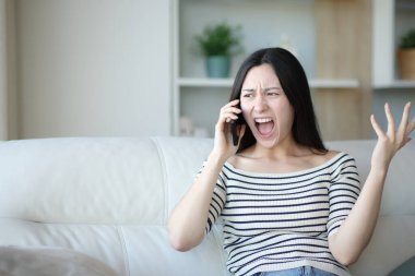 Kızgın Asyalı kadın telefonda konuşuyor. Evdeki kanepede oturuyor.