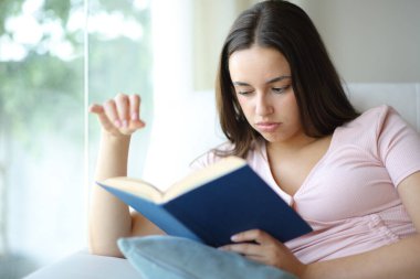 Evde bir kanepede oturup kötü bir kağıt kitap okuyan sıkıcı bir kadın.