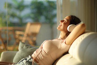 Mutlu kadın, gün batımında evdeki bir kanepede oturan, kablosuz kulaklıktan sesi dinliyor.