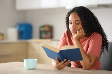 Rahatlamış, ciddi, siyah bir kadın mutfakta oturmuş kitap okuyor.