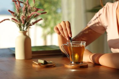 Gün batımında, bir evin içinde, ahşap masanın üstünde çay karıştıran bir kadının elini yakın plan.