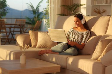 Gün batımında kulaklıklı mutlu bir kadın dizüstü bilgisayar kullanarak evdeki koltukta oturuyor.