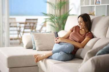 Oturma odasındaki kanepede oturmuş akıllı telefon kullanan mutlu bir kadın.