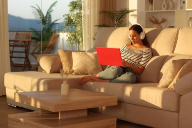 Kulaklık takan, kırmızı dizüstü bilgisayar kullanan bir kadın gün batımında oturma odasındaki koltukta oturuyor.