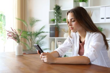 Evdeki ahşap masada oturan rahat bir kadın cep telefonu kullanıyor.