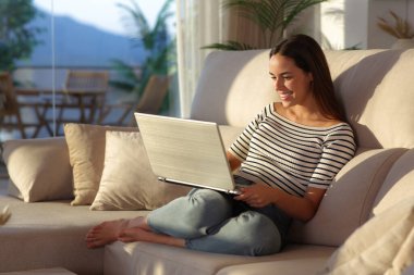 Evdeki koltukta oturup dizüstü bilgisayar kullanan mutlu bir kadın.