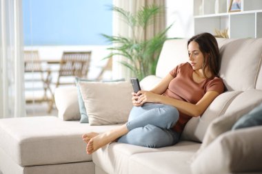 Evin içindeki bir koltukta oturan ciddi bir kadın cep telefonu kullanıyor.