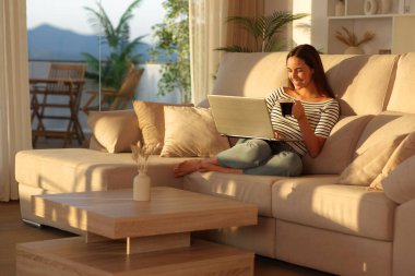 Evdeki bir kanepede oturup laptopunu kullanan mutlu kadın kahve içiyor.