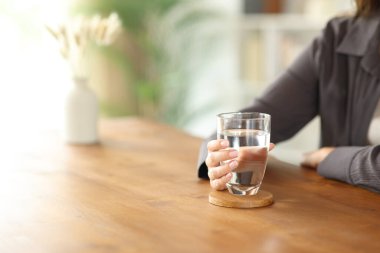 Evdeki ahşap masada su bardağı tutan bir kadının elini yakın plan.