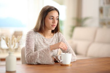 Kafası karışık bir kadın kahvesini karıştırıyor. Evdeki oturma odasına bakıyor.