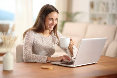 Evdeki bir oturma odasında dizüstü bilgisayar kullanan ve kahve içen mutlu bir kadın.