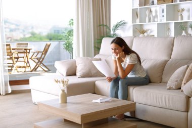 Evdeki kanepede oturup kağıt okuyan mutlu bir kadın.