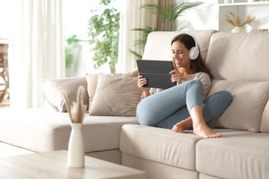 Evdeki kanepede oturup video izlemek için tablet ve kablosuz kulaklık kullanan mutlu kadın.