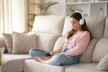 Evde telefon dinleyen bir kadın var. Kulaklıkla koltukta oturuyor.