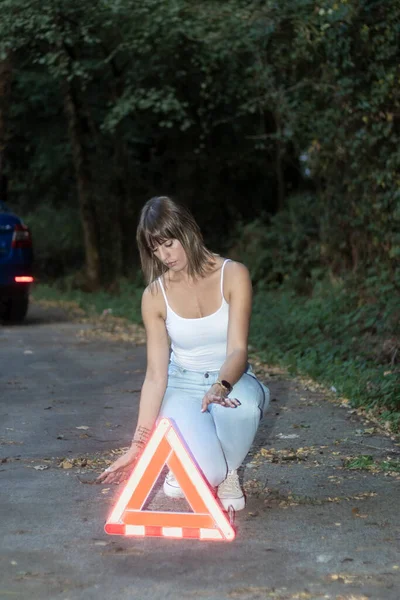 Young woman putting a caution sign in the red triangle on the road near a broken car on the road.