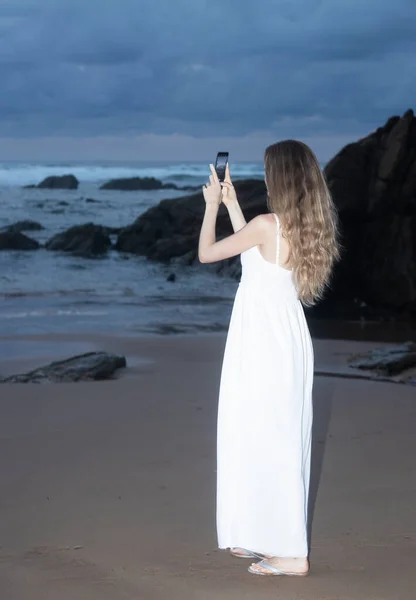 Woman holding mobile phone at sunset. Young woman taking photos with her cell phone in a beautiful amazing sunset over sea. Taking a picture on a smartphone during a vacation
