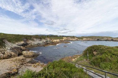 Mavi gökyüzü, beyaz bulutlar ve küçük bir sahil bölgesi olan kayalık kıyı şeridi. Ön planda metal bir korkuluk görünüyor.