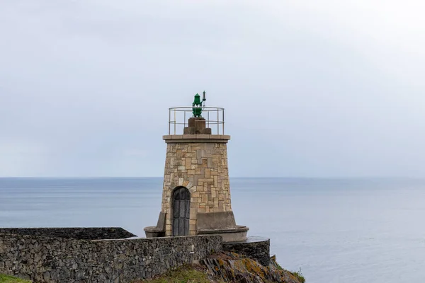 stone lighthouse with a green statue on top, overlooking the calm ocean under an overcast sky