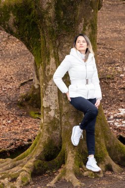 Ormanda yosun kaplı bir ağaç gövdesinde oturan kadın, beyaz ceket ve siyah pantolon giyiyor.