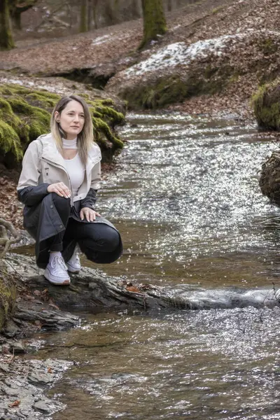 woman sitting on a rock by a stream in a forest, wearing a white jacket, black pants, and white shoes